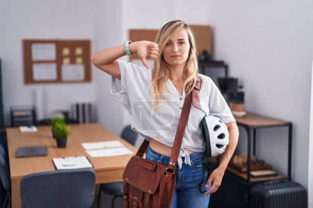 Photo for Young blonde woman working at the office holding bike helmet with angry face, negative sign showing dislike with thumbs down, rejection concept - Royalty Free Image