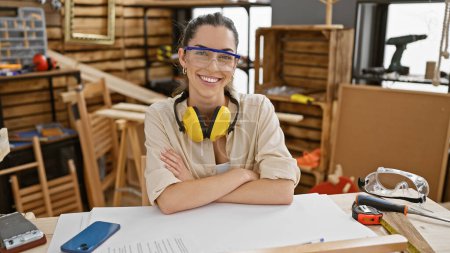Photo for Young beautiful hispanic woman carpenter wearing glasses sitting on table with arms crossed gesture at carpentry - Royalty Free Image