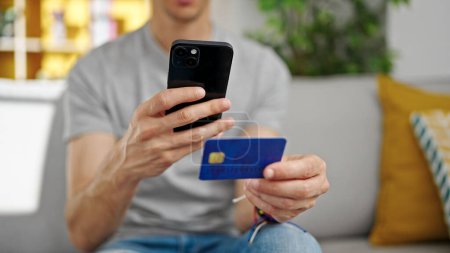 Téléchargez les photos : Jeune homme hispanique faire une photo à la carte de crédit par smartphone à la maison - en image libre de droit