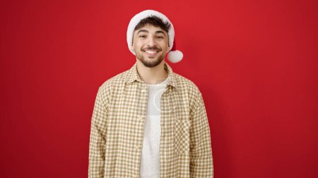 Foto de Hombre árabe joven sonriendo confiado usando sombrero de Navidad sobre fondo rojo aislado - Imagen libre de derechos
