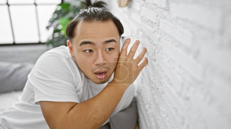 Photo for Nosy young chinese man sneakily listening through bedroom wall with glass, overcome by curiosity and eavesdropping in a secret indoor spy mission in his apartment. - Royalty Free Image