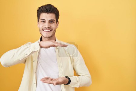 Téléchargez les photos : Young hispanic man standing over yellow background gesturing with hands showing big and large size sign, measure symbol. smiling looking at the camera. measuring concept. - en image libre de droit