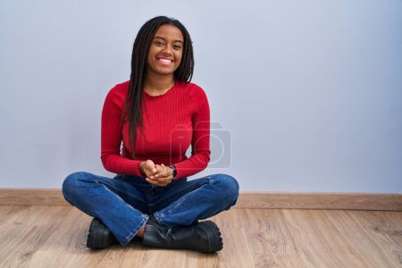 Foto de Joven afroamericano con trenzas sentado en el suelo en casa con las manos juntas y los dedos cruzados sonriendo relajado y alegre. éxito y optimismo - Imagen libre de derechos