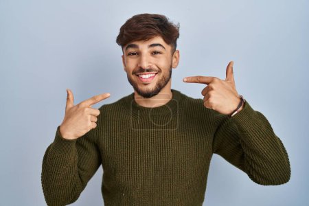 Téléchargez les photos : Homme arabe avec barbe debout sur fond bleu souriant gai montrant et pointant avec les doigts dents et la bouche. concept de santé dentaire. - en image libre de droit