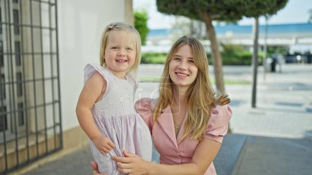 Photo for Cheerful caucasian mother and daughter embracing a warm, loving hug, sitting on a city bench, laughing, and smiling together, filled with joy and happiness, outdoors on a sunny street. - Royalty Free Image
