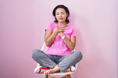Photo for Hispanic young woman sitting on chair over pink background smiling with hands on chest with closed eyes and grateful gesture on face. health concept. - Royalty Free Image