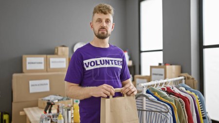 Un beau jeune homme avec une barbe, vêtu d'une chemise volontaire, se tient fièrement dans un centre de dons avec des boîtes et des vêtements en arrière-plan.