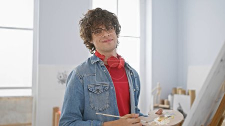 Foto de Joven confiado con gafas y auriculares está en un estudio de arte, sosteniendo una paleta de pintura y un pincel. - Imagen libre de derechos