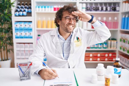 Photo for Hispanic young man working at pharmacy drugstore very happy and smiling looking far away with hand over head. searching concept. - Royalty Free Image
