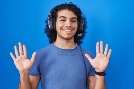 Téléchargez les photos : Homme hispanique aux cheveux bouclés écoutant de la musique à l'aide d'écouteurs montrant et pointant vers le haut avec les doigts numéro dix tout en souriant confiant et heureux. - en image libre de droit