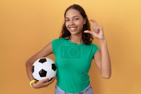Téléchargez les photos : Jeune femme hispanique tenant balle geste souriant et confiant avec la main faisant signe de petite taille avec les doigts regardant et la caméra. concept de mesure. - en image libre de droit
