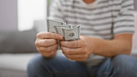 Photo for Handsome young caucasian man counting dollars on sofa at home, managing personal finance in the living room - Royalty Free Image