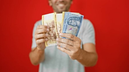 Téléchargez les photos : Jeune homme souriant, ravi de compter sa richesse en billets de cent forints sur fond rouge isolé - en image libre de droit