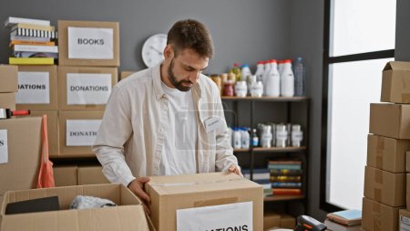 Photo for Altruistic young hispanic man volunteering at charity center, concentrated on holding a package of donations amidst community work in a storehouse - Royalty Free Image