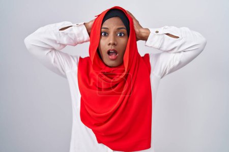 Téléchargez les photos : Jeune femme arabe portant l'écharpe traditionnelle islamique hijab fou et effrayé avec les mains sur la tête, effrayé et surpris de choc avec la bouche ouverte - en image libre de droit