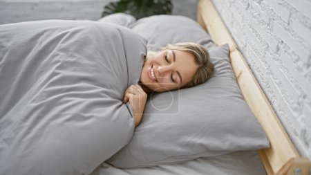 Photo for Blonde young woman smiling peacefully while lying in bed indoors, portraying comfort and relaxation at home. - Royalty Free Image