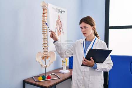 Téléchargez les photos : Jeune femme blonde pysiothérapeute pointant vers le modèle anatomique de colonne vertébrale tenant presse-papiers à la clinique de réadaptation - en image libre de droit