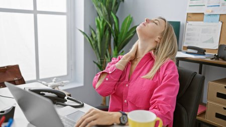 Foto de Una joven rubia con una camisa rosa sintiendo dolor de cuello en el escritorio de su oficina, exudando una mezcla de profesionalismo e incomodidad. - Imagen libre de derechos