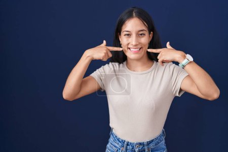 Photo for Young hispanic woman standing over blue background smiling cheerful showing and pointing with fingers teeth and mouth. dental health concept. - Royalty Free Image