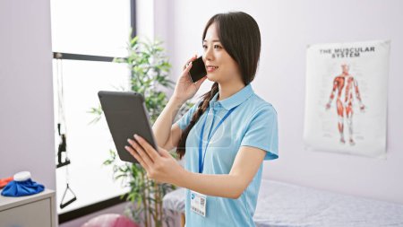 Téléchargez les photos : Une jeune femme asiatique professionnelle de la santé parle sur un téléphone tout en tenant une tablette dans un intérieur de clinique. - en image libre de droit