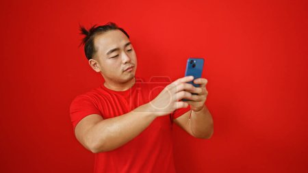 Téléchargez les photos : Jeune homme chinois grossier plongé dans une conversation en ligne dynamique sur son smartphone, isolé sur un fond rouge audacieux. - en image libre de droit
