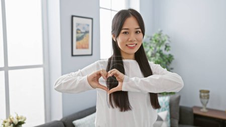 Téléchargez les photos : Une jeune femme asiatique dans un chemisier blanc créant une forme de coeur avec ses mains dans un salon lumineux. - en image libre de droit