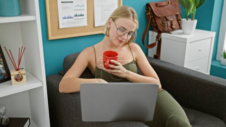 Foto de Joven mujer rubia trabajadora de negocios usando laptop bebiendo café en la oficina - Imagen libre de derechos