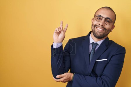 Foto de Hombre hispano con barba vistiendo traje y corbata sonriendo con la cara feliz guiñando a la cámara haciendo señal de victoria. número dos. - Imagen libre de derechos