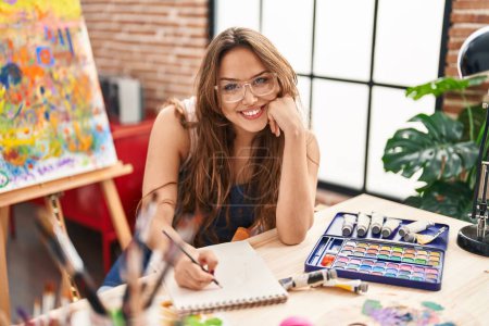 Téléchargez les photos : Jeune belle artiste hispanique femme souriante dessin confiant sur carnet au studio d'art - en image libre de droit