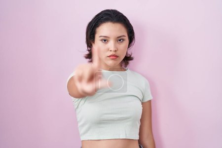 Foto de Mujer joven hispana de pie sobre fondo rosa señalando con el dedo hacia arriba y la expresión enojada, sin mostrar ningún gesto - Imagen libre de derechos