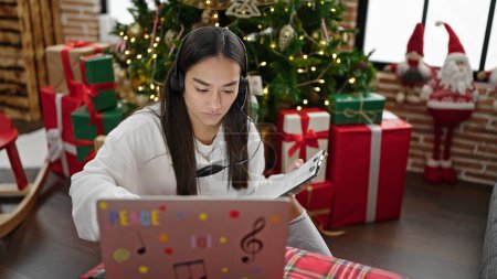 Photo for Young beautiful hispanic woman using laptop and headphones writing on document at home - Royalty Free Image