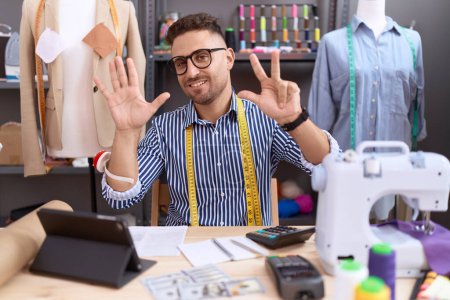 Photo for Hispanic man with beard dressmaker designer working at atelier showing and pointing up with fingers number eight while smiling confident and happy. - Royalty Free Image