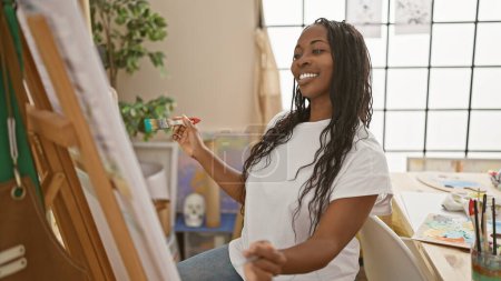 Téléchargez les photos : Femme afro-américaine souriante peignant sur une toile dans un studio d'art lumineux - en image libre de droit