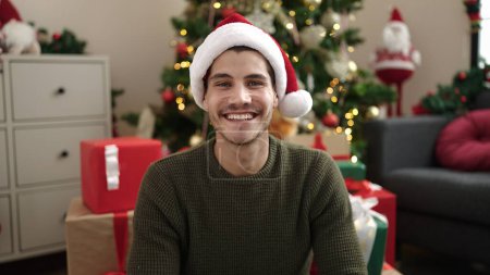Foto de Joven hombre hispano sonriendo confiado sentado en el suelo por el árbol de Navidad en casa - Imagen libre de derechos