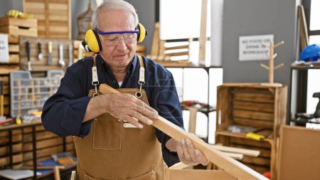 Téléchargez les photos : L'homme âgé résilient, charpentier chevronné, tient ferme dans son atelier de menuiserie, enfilant des lunettes de sécurité et tenant une planche de bois fraîchement sciée. - en image libre de droit