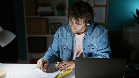 Téléchargez les photos : Un jeune homme concentré dans une veste en jean écrit sur un document à son bureau à domicile pendant la nuit. - en image libre de droit
