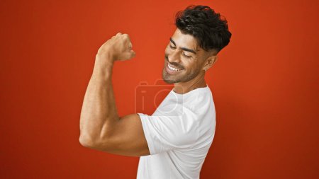 Photo for Handsome young hispanic man flexing muscles with a smile against a red wall background - Royalty Free Image