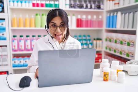 Téléchargez les photos : Jeune femme arabe travaillant à la pharmacie pharmacie en utilisant un ordinateur portable collant la langue heureux avec une expression drôle. concept d'émotion. - en image libre de droit