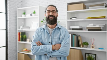 Photo for A confident hispanic man with a beard standing in a modern office setting, arms crossed and smiling. - Royalty Free Image