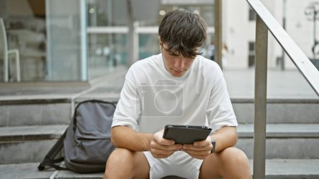Foto de Joven adolescente hispano fresco, un estudiante serio, sentado casualmente en las escaleras de la universidad, absorto en la pantalla de su computadora portátil - concentrado en el touchpad, encarnando el estilo de vida de tecnología inteligente al aire libre. - Imagen libre de derechos