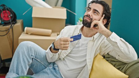 Téléchargez les photos : Jeune homme hispanique tenant une carte de crédit assis sur le canapé pensant à la nouvelle maison - en image libre de droit