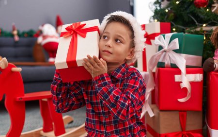 Photo for Adorable hispanic boy hearing gift sound sitting on floor by christmas tree at home - Royalty Free Image