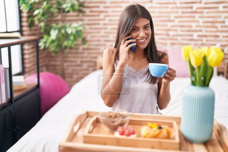 Photo for Young beautiful hispanic woman having gift breakfast talking on smartphone at bedroom - Royalty Free Image