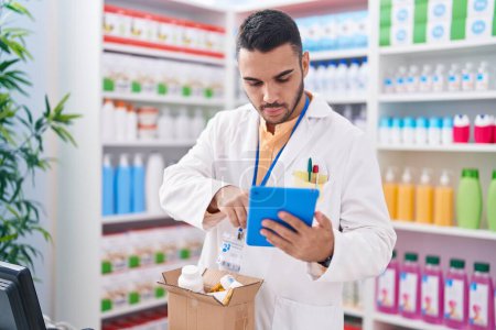 Téléchargez les photos : Jeune homme hispanique pharmacien en utilisant touchpad tenant bouteille pilules à la pharmacie - en image libre de droit