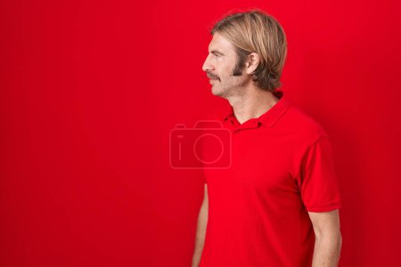 Téléchargez les photos : Homme caucasien avec moustache debout sur fond rouge regardant vers le côté, pose de profil relax avec visage naturel avec sourire confiant. - en image libre de droit