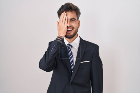 Téléchargez les photos : Jeune homme hispanique avec des tatouages portant un costume d'affaires et cravate couvrant un oeil avec la main, sourire confiant sur le visage et l'émotion surprise. - en image libre de droit