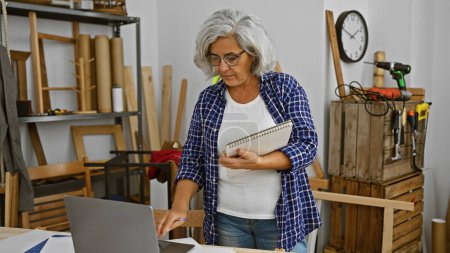 Photo for Mature woman reviews plans in a woodworking workshop surrounded by tools and lumber - Royalty Free Image