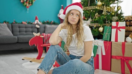 Photo for Young blonde woman celebrating christmas sitting on floor with sad expression at home - Royalty Free Image
