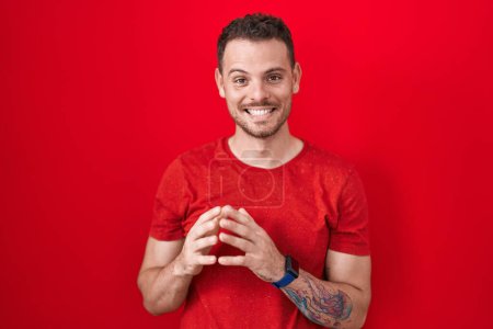 Téléchargez les photos : Jeune homme hispanique debout sur fond rouge mains ensemble et doigts croisés souriant détendu et joyeux. succès et optimisme - en image libre de droit