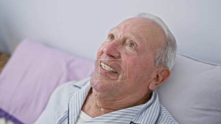 Téléchargez les photos : Homme âgé confiant, avec un sourire chaleureux, profitant d'une matinée confortable couché dans le lit dans l'atmosphère intime de sa chambre confortable. - en image libre de droit